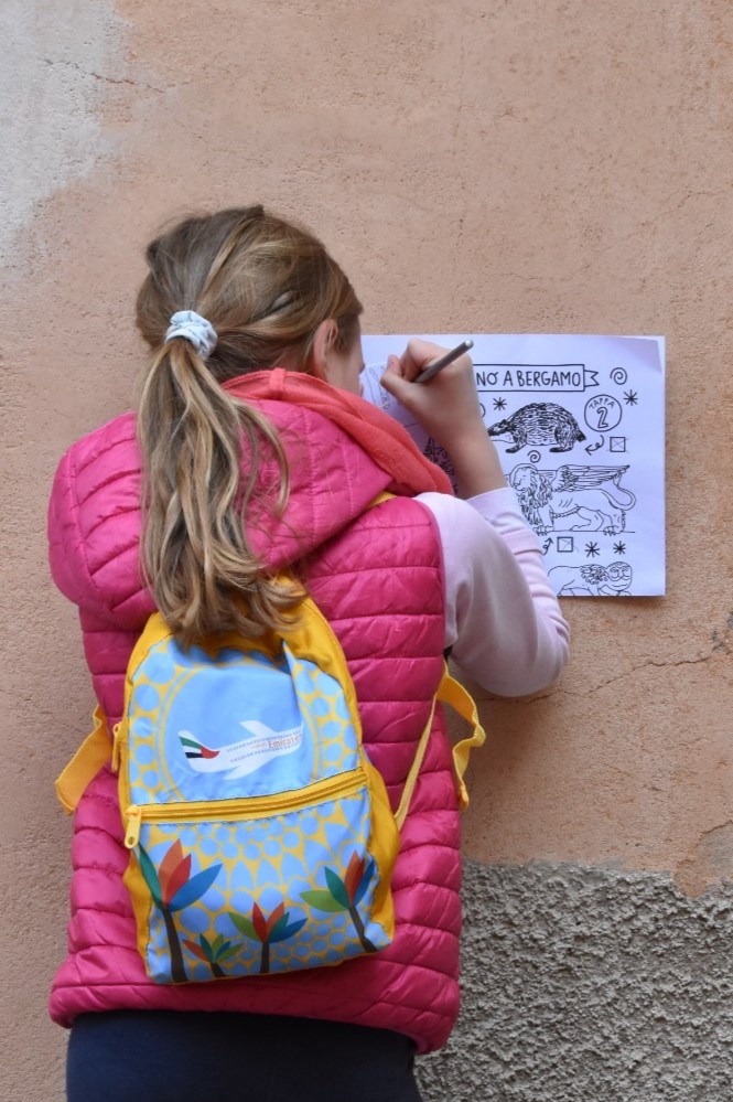 Una bambina sta scrivendo sulla mappa illustrata del Safari Urbano di Bergamo, appoggiandosi al muro di una via di Città Alta.